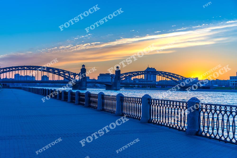 На фотографии изображён Большеохтинский мост, подсвеченный мягким светом белых ночей. На переднем плане расположена набережная Невы с изящными чугунными ограждениями, создающими элегантную рамку для вида на мост и реку. Лёгкое свечение на небе подчёркивает уникальность этого явления, характерного для Петербурга летом. Большеохтинский мост остаётся одним из символов инженерного мастерства и красоты города.